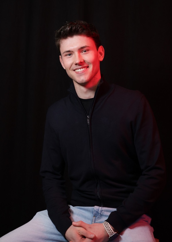Tomáš Klíma, smiling, in a black zip-up against a dark backdrop with red side light